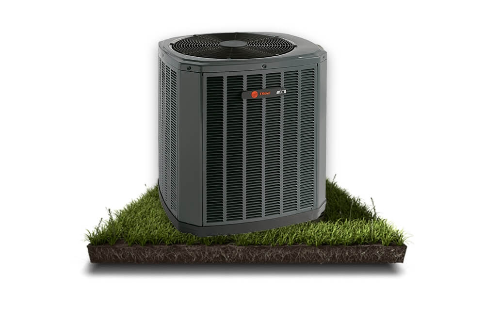 A black air conditioner sitting on a patch of green grass with a white background. The air conditioner has a metal cabinet with a fan on the top and a vent on the front. There are electrical wires and pipes connected to the back of the air conditioner.